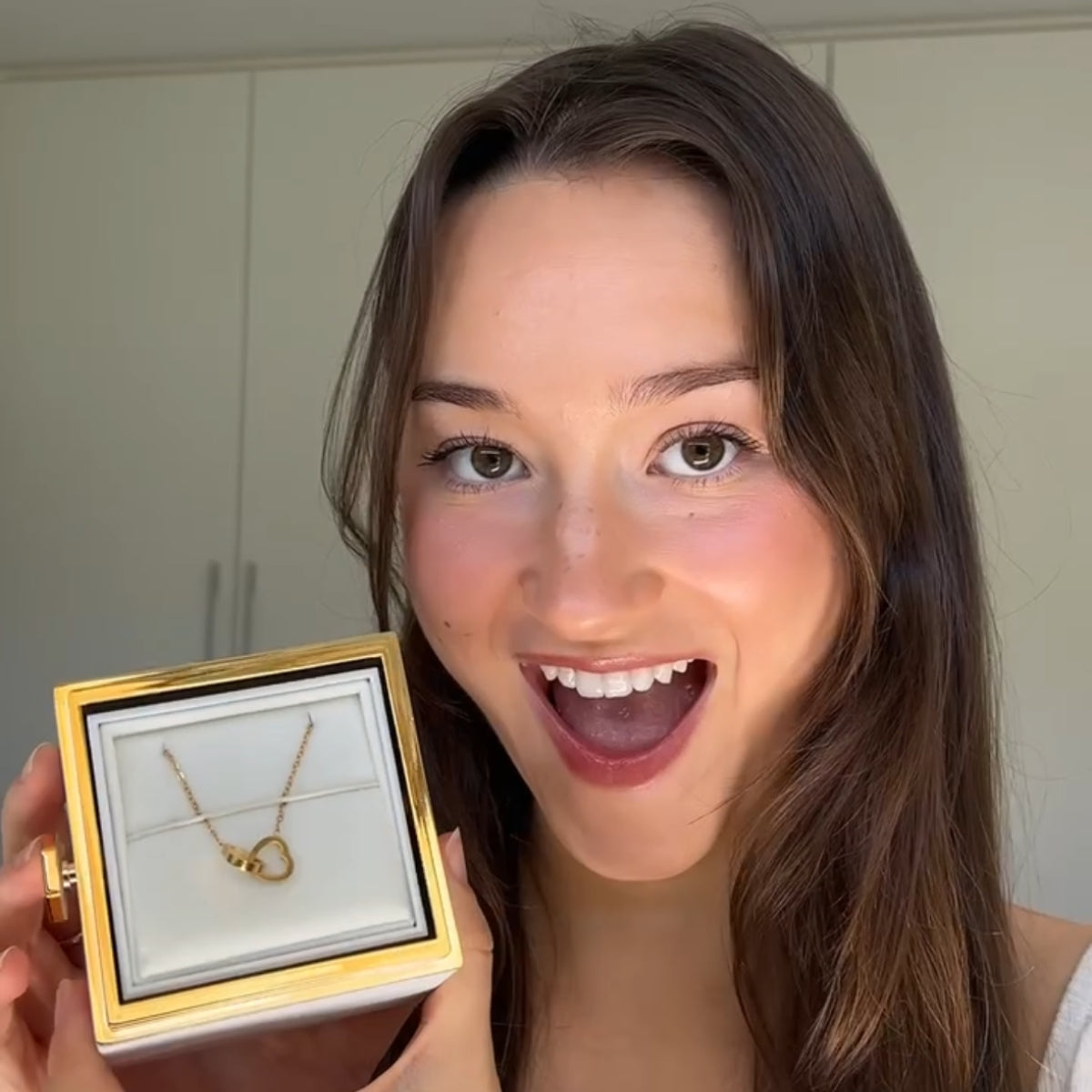 Woman smiling while holding a gold-trimmed box displaying the gold double-heart necklace.